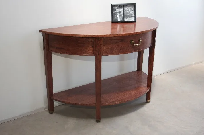 A half-round cherry table with a drawer and a lower shelf, standing against a white wall