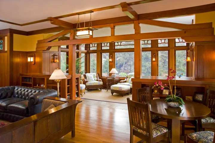 An open living and dining room framed by exposed fir posts and beams, with a hanging lantern, a leather sofa and a wall of wood-trimmed windows