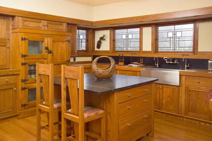 A kitchen with wood cabinets, a dark stone island with two wooden chairs, and leaded-glass windows over the sink