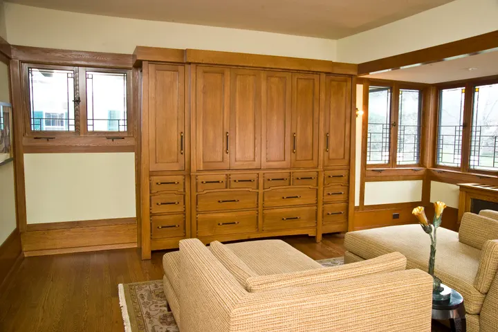 A wall-to-wall oak wardrobe with tall cabinet doors over rows of drawers, set between two windows, with a cream sofa in front
