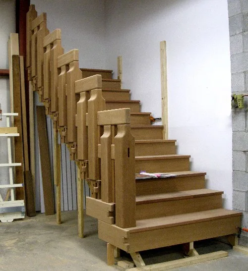 An unfinished wooden staircase with tall square posts and stepped rails, standing on the floor of a workshop