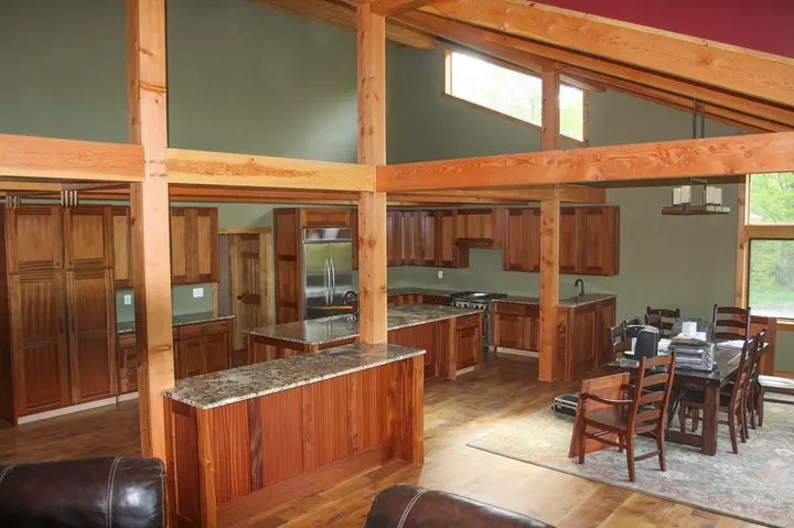 An open kitchen under exposed fir posts and beams, with wood cabinets, a granite-topped island and a dining table beyond
